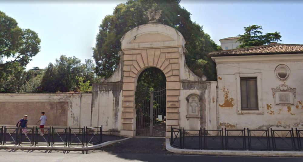 Historic arched gate with stone sculptures, adjacent to an old building; two people walking nearby.