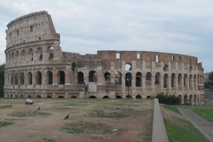 Side view of the ancient Colosseum in Rome with a grassy foreground and cloudy sky.