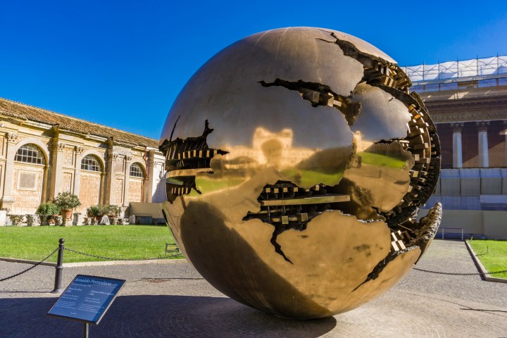 Bronze globe sculpture with cracks, set in a courtyard with blue sky.