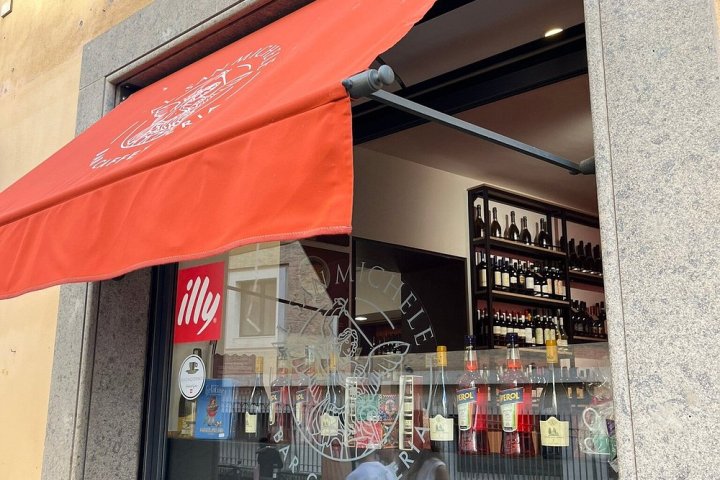 Cafe with red awning, people seated, bottles and logo on window, outdoor table.